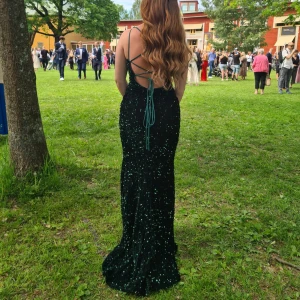 Balklänning  - Säljer min mörkgrön långklänning med glittriga paljetter och djup v-ringning. Klänningen har smala axelband, snörning i ryggen och en slits framtill. Perfekt för bal eller student! Snygg och figurnära passform som ger en elegant siluett. 