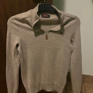 Beige merinoullströja från Morris - Snygg beige långärmad tröja från Morris i 100% merinoull. Tröjan har en hög krage med half zip-dragkedja och en liten broderad logga på bröstet. Perfekt för lager-på-lager och stilrena outfits. Passar dig som är 165-170cm.      PRISET ÄR FAST då tröjan fortfarande är i ett mycket bra skick