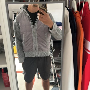 Grå quiltad cardigan - Säljer en grå cardigan med stickade ärmar och krage. Cardiganen har quiltad front i mörkgrått, två fickor och dragkedja. Snygg mix av material med stickad struktur på ärmar och sidor, perfekt för lager-på-lager och chill stil.