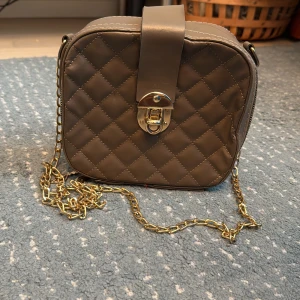 Beige.brun  quiltad axelväska med kedja - Snygg beige/brun  axelväska med quiltat mönster och guldfärgad kedja som axelrem. Väskan har en rundad fyrkantig form, dekorativ låsdetalj i metall och dragkedja på sidan. Perfekt för att lyfta din outfit med lite extra stil.