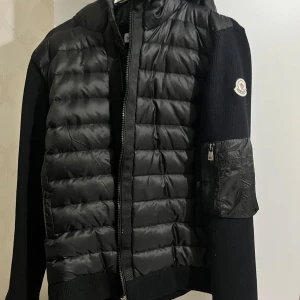 Svart Moncler pufferjacka med huva - Svart pufferjacka från Moncler med stickade ärmar och quiltad front. Jackan har huva, dragkedja och en snygg ficka med zip på ärmen. Ikonisk Moncler-logga på vänster ärm. Perfekt för dig som vill ha en clean och exklusiv look.
