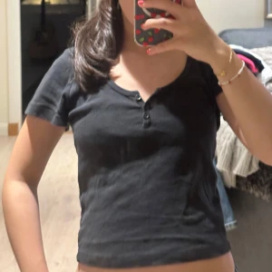 Svart croppad topp med knappar - Säljer en svart croppad topp med korta ärmar och rundad halsringning. Toppen har tre små knappar framtill som ger en chill och enkel vibe. Perfekt att matcha med jeans eller kjol för en avslappnad look. Från brandy!!