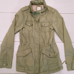 Olivgrön Höstjacka - En olivgrön höstjacka från MQ, storlek XS💕 sann i storlek. Har en massa coola detaljer såsom störning i midjan så att den går att göra tightare eller en ficka vid nacken vars man kan ta fram en luva⭐ Jackan har använts sparsamt och har inga hål eller liknande! 