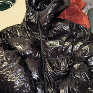 Svart pufferjacka från DSQUARED2 - Säljer en svart, glansig pufferjacka från DSQUARED2 med huva och dragkedja framtill. Jackan har en fet DSQ2-logga på bröstet och är tillverkad i polyester. Perfekt för dig som vill ha en cool och varm jacka med streetkänsla.
