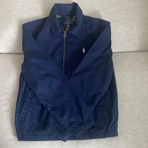 Marinblå vindjacka från Polo Ralph Lauren - Snygg marinblå vindjacka från Polo Ralph Lauren med klassisk krage och vit broderad logga på bröstet. Jackan har dragkedja framtill, två sidofickor och elastiska muddar vid ärmslut och nederkant. Perfekt för dig som gillar stilren och sportig look och den är helt oanvänd