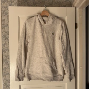 Ljusgrå hoodie från Lyle & Scott - Säljer en ljusgrå hoodie från Lyle & Scott Junior med klassisk gul broderad logga på bröstet. Tröjan har huva, magficka och långa ärmar. Perfekt för chill dagar och enkel att matcha med jeans eller joggers.