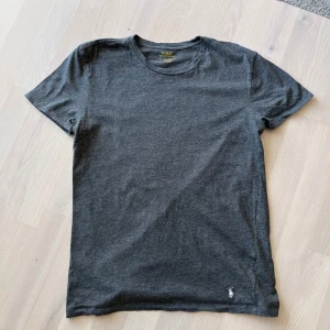 Mörkgrå t-shirt från Polo Ralph Lauren - Snygg mörkgrå t-shirt från Polo Ralph Lauren med klassisk rund hals och korta ärmar. Tillverkad i mjuk bomull och har den ikoniska lilla Polo-loggan broderad i vitt nedtill. Perfekt basic för garderoben.