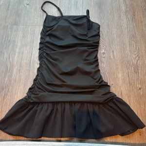 Svart volangklänning från SHEIN - Svart kortklänning från SHEIN med smala axelband och snygga rynkade detaljer längs sidorna. Nederdelen har en bred volang som ger extra flärd. Klänningen är tillverkad i polyester med stretchig elastan för en tight och bekväm passform. Perfekt för dig som gillar trendiga och stilrena plagg.