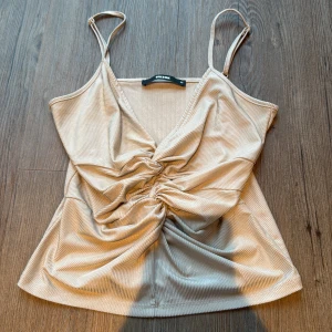 Beige ribbad linnetopp från BikBok - Snygg beige linnetopp från BikBok i ribbad syntetmaterial. Toppen har smala axelband och en twistad detalj framtill som ger en cool och trendig look. Perfekt för varma dagar och passar till både jeans och kjol. Färgen är ljusbeige och materialet har en lätt glans.
