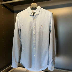 Blå skjorta från Polo Ralph Lauren - Snygg ljusblå skjorta från Polo Ralph Lauren i slim fit. Klassisk button-down krage, broderad logga på bröstet och vita knappar framtill. Tillverkad i mjuk bomull och har långa ärmar med manschettknappar. Perfekt för dig som gillar stilrena plagg.