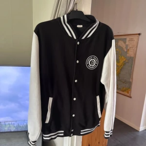 Svart och vit collagejacka med tryck - Snygg svart och vit bomberjacka med klassisk college-look. Jackan har vita ärmar, svart kropp och vita detaljer som knappar och fickor. Ribbad krage och muddar med svarta och vita ränder. Tryck på bröstet med texten 'Kunskap förändrar'. Perfekt för dig som gillar streetstyle.