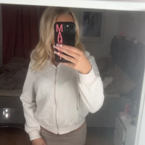 Vit hoodiejacka med dragkedja - Säljer en vit hoodiejacka med dragkedja och huva. Jackan har ribbade muddar vid ärmslut och nederkant samt snörning i huvan. Perfekt för chill dagar och enkel att matcha med jeans eller kjol. Mjuk och skön bomullskänsla.