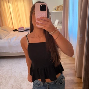 Svart linnetopp med volangkant - Säljer en svart linnetopp med smala axelband som knyts på axlarna. Toppen har en rak halsringning och en söt volangkant nedtill som ger en chill och feminin vibe. Perfekt att matcha med jeans eller kjol för en enkel och snygg look.