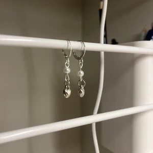 Silvriga örhängen med pärlor - Snygga silvriga örhängen med hängande detaljer. Varje örhänge har en liten vit pärla och ett hjärt hänge i silverfärg. Perfekt för dig som gillar stilrena och trendiga accessoarer. I rostfritt stål och handgjort 