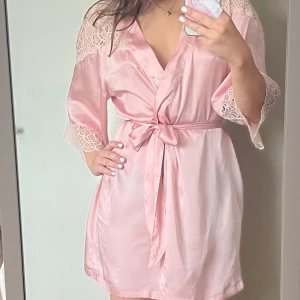 Rosa satinmorgonrock med spetsdetaljer - Säljer en ljusrosa kort morgonrock i satin. Klänningen har  spetsdetaljer på axlar och ärmslut samt trekvartsärmar. Den är från hm men en gammal kollektion, lappen sitter tyvärr inte kvar längre då den skavde. Ett hål i tyget, men det syns ej när man har den på sig. Se bild 4.