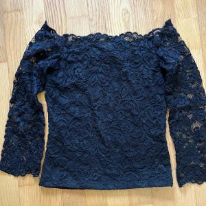 Svart offshoulder spetsblus - Supersnygg svart blus med offshoulder och långa ärmar i genombruten spets. Blommigt spetsmönster över hela toppen ger en romantisk vibe. Perfekt att styla med jeans eller kjol för en trendig look.