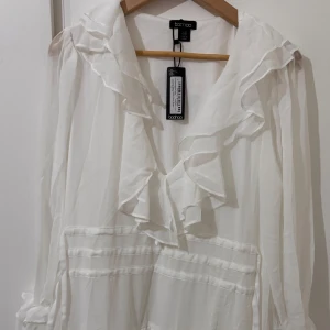 Vit volangklänning från Boohoo - Supersöt vit klänning från Boohoo med massor av volanger och romantiska detaljer. Klänningen har lång ärm, v-ringad hals och flera lager volanger både på ärmar och nederdel. Perfekt för dig som gillar en feminin och flowy stil. Passar perfekt för studenten!