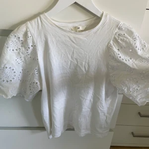 Vit blus från H&M - Säljer en söt vit blus från H&M med puffiga kortärmar i broderad spets. Blusen har en rund halsringning och är tillverkad i ett mjukt bomullsmaterial.