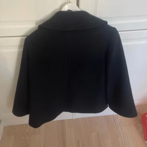 Svart kappa från Yera - Elegant svart kappa från Yera i storlek M. Den har en klassisk design med knappar framtill och en bred krage. Perfekt för kyligare dagar.