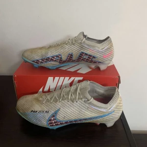 Nike Air Zoom fotbollsskor - Snygga Nike Air Zoom fotbollsskor i beige med blå och rosa detaljer. Skorna har snörning och en rund tå, perfekt för fotbollsplanen. De har en modern design med ett mönster som ger extra stilpoäng.