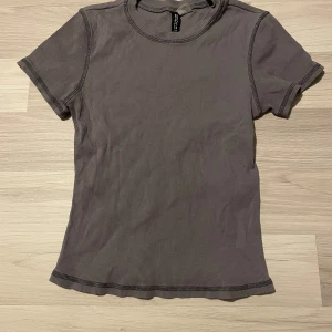 Grå ribbad t-shirt från H&M - Snygg grå ribbad t-shirt från H&M. Den har en tight passform och korta ärmar, perfekt för en stilren look. T-shirten är gjord i ett mjukt material som känns skönt mot huden.