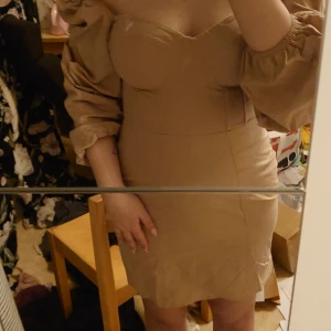 Beige klänning med korset detalj - Säljer en stilren beige klänning med långa puffärmar och en figurnära passform. Klänningen har en fyrkantig urringning och dragkedja i ryggen. Perfekt för en elegant look! Man kan justera ärmarna och ha en mer scrunchie look om man vill. Dragkedja går upp hela vägen det var bara svårt att göra själv så det är därför den inte är hela vägen upp i bilden. 