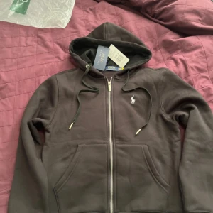 Svart hoodie från Polo Ralph Lauren - Säljer en stilren svart hoodie från Polo Ralph Lauren med dragkedja och huva. Den har en broderad logga på bröstet och praktiska fickor framtill. Perfekt för en avslappnad look.