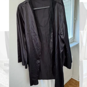 Svart lång satinrock - Elegant svart lång kappa med en öppen design. Perfekt för en stilren look. Den har långa ärmar och ett mjukt, glansigt material som ger en lyxig känsla.