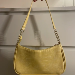 Gul axelväska med krokodilmönster - Snygg gul axelväska med krokodilmönster och dragkedja. Väskan har en kedjedetalj på axelremmen och en rymlig insida. Perfekt för att ge en färgklick till din outfit!