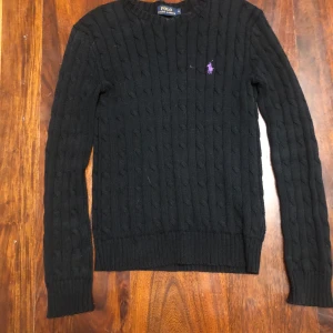 Svart stickad tröja från Ralph Lauren - Säljer en klassisk svart stickad tröja från Ralph Lauren med kabelstickat mönster och lila logotyp på bröstet. Tröjan har långa ärmar och ribbstickade muddar. Perfekt för en stilren look.