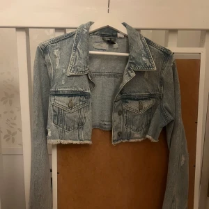 Ljusblå croppad jeansjacka - Säljer en ljusblå croppad jeansjacka med slitna detaljer och fransad nederkant. Jackan har långa ärmar och två bröstfickor med knappar. Perfekt för en avslappnad stil!