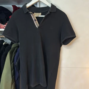 Svart kortärmad t-shirt från Burberry - Snygg svart kortärmad t-shirt från Burberry med klassisk krage och knappdetalj i det ikoniska Burberry-mönstret. Perfekt för en stilren look.