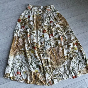 Mönstrad kjol från H&M - Säljer en mönstrad kjol från H&M i storlek XS. Kjolen har ett tropiskt mönster med gröna och bruna nyanser samt knappar framtill. Perfekt för en somrig look!
