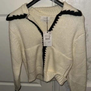 Vit stickad kofta från Zara - Säljer en vit stickad kofta från Zara med svarta detaljer längs dragkedjan och kragen. Koftan har en klassisk krage och är perfekt för kyligare dagar. Passar både till vardags och lite finare tillfällen.