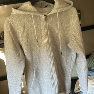 Grå hoodie från Jack & Jones - Säljer en stilren grå hoodie från Jack & Jones. Den har en dragkedja framtill och justerbar huva med snören. Perfekt för en avslappnad look.