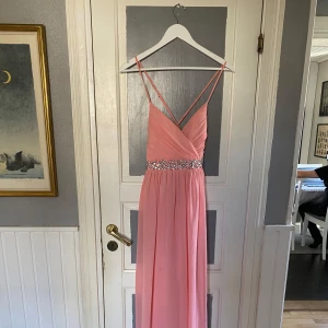 Rosa balklänning med diamantdetaljer  - Elegant rosa långklänning med smala axelband och omlott-design. Klänningen har en vacker strassdetalj i midjan och en öppen rygg som ger en sofistikerad look. Den har dock smutsrken på ryggen. Därav sänkt pris 