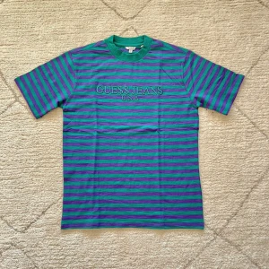 Randig t-shirt från Guess - Säljer en cool randig t-shirt från Guess i grönt och lila med 'GUESS JEANS U.S.A' broderat på bröstet. T-shirten har en normal passform och korta ärmar, perfekt för en avslappnad stil.