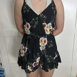 Svart blommig jumpsuit - Säljer en svart jumpsuit med vackert blommönster i rosa och grönt. Den har smala axelband och en v-ringad design. Perfekt för sommarens varma dagar!