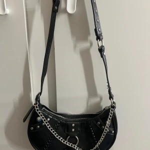 Svart axelväska med kedjedetalj - Snygg svart axelväska med justerbar rem och kedjedetalj. Väskan har en dragkedja upptill och dekorativa nitar. Perfekt för att ge din outfit en edgy touch. Själva dragkedjan har lossnat för fortfarande att stänga