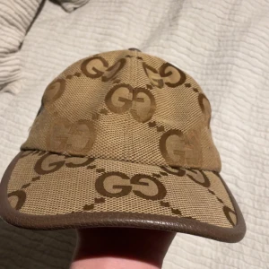 Beige keps från Gucci - Snygg beige keps från Gucci med brunt GG-mönster. Kepsen har en justerbar rem baktill och är kantad med brunt läder. Perfekt för att ge din outfit en lyxig touch.Vid en snabb affär kan jag gå ned i pris.