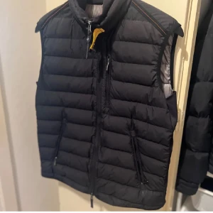 Svart dunväst från Parajumpers - Säljer en svart dunväst från Parajumpers i superlätt design. Västen har dragkedja och är perfekt för lager-på-lager under kyliga dagar. Den har en stilren look med quiltad yta och en liten gul detalj vid kragen. Inga defekter förutom den lila hålet i bild nr 5