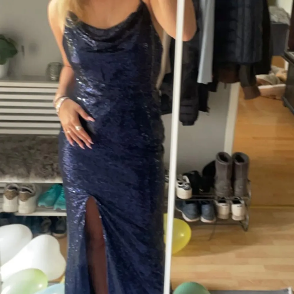 Snygg mörkblå klänning med glittrande paljetter och en elegant slits framtill. Klänningen har en draperad halslinje och är ärmlös, perfekt för festliga tillfällen som till exempel bal . Har en lite mer öppen rygg med korsande band . Mekot.