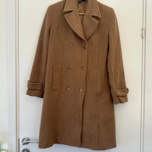 Beige kappa från KappAhl - Säljer en stilren beige kappa från KappAhl med klassisk krage och knappar framtill. Kappan har långa ärmar med dekorativa knappar vid ärmsluten. Perfekt för höst och vår!