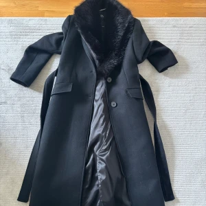 Svart kappa från Zara med fuskpäls - Elegant svart kappa från Zara med fuskpälsdetaljer på kragen. Kappan har en klassisk design med knappar framtill och ett bälte i midjan för en smickrande passform. Perfekt för kyligare dagar.
