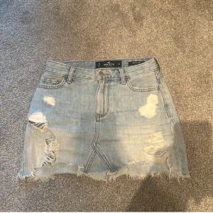 Jeanskjol från Hollister - Snygg jeanskjol från Hollister med slitna detaljer och fransig nederkant. Kjolen har en klassisk femficksdesign och en ljusblå tvätt. 