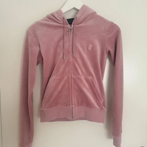 Rosa velour hoodie från Juicy Couture - Säljer en mjuk och stilren rosa velour hoodie från Juicy Couture. Den har dragkedja framtill, justerbar huva med snören och två fickor. Perfekt för en avslappnad look.