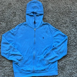 Blå hoodie från Polo Ralph Lauren - Säljer en snygg blå hoodie från Polo Ralph Lauren med dragkedja och klassisk logga på bröstet. Perfekt för en avslappnad stil. Hoodien har långa ärmar och en bekväm passform.