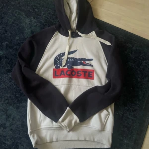 Svart och vit hoodie från Lacoste - Säljer en snygg svart och vit hoodie från Lacoste med det klassiska krokodilmärket och texten 'Lacoste' i rött på framsidan. Hoodien har en justerbar dragsko i huvan och långa ärmar. Perfekt för en avslappnad stil. Aldrig använd.