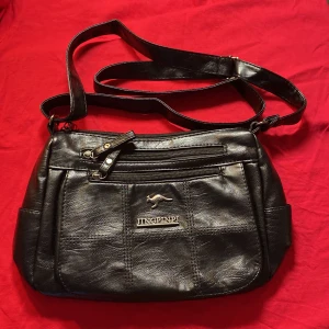 Svart axelväska från JINGPINP - Helt ny .Snygg svart axelväska i skinn från JINGPINP med flera praktiska fack och dragkedjor. Väskan har en justerbar axelrem och en stilren design med en logotyp framtill. Perfekt för att bära med sig det nödvändigaste. 