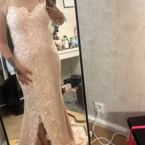 Champagnefärgad spetsklänning - Elegant långklänning i champagnefärgad spets med vackra broderier och en subtil slits framtill. Klänningen är axelbandslös och har en figurnära passform som framhäver silhuetten. Perfekt för speciella tillfällen.